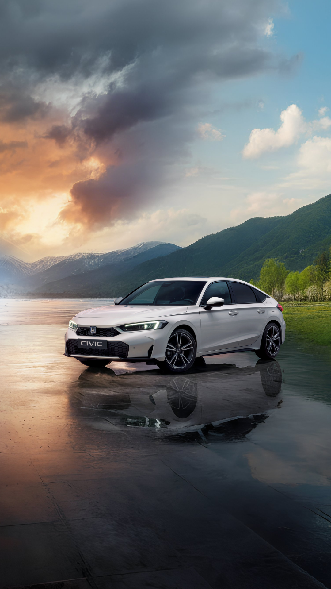A silver Honda Civic sedan parked on a reflective surface near a lake, with mountains and a dramatic sky in the background. The sky features a mix of storm clouds and sunset colors, creating a striking contrast with the car's sleek design.